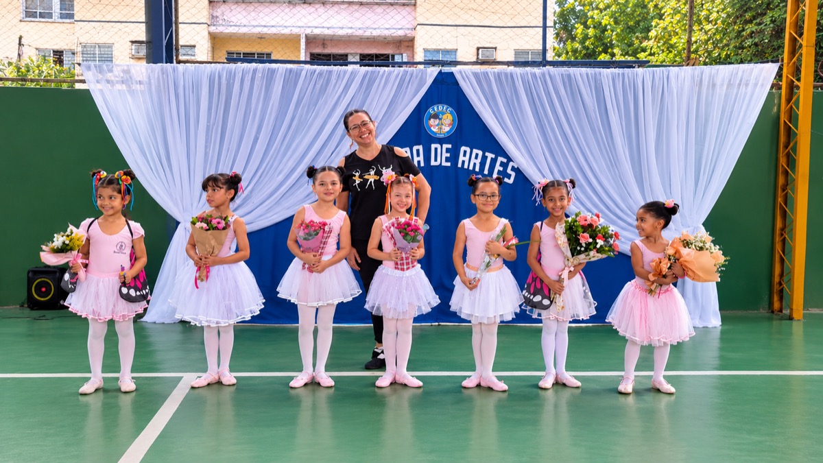 Alunas do balé com a professora, de collant rosa e saia de tule, segurando flores